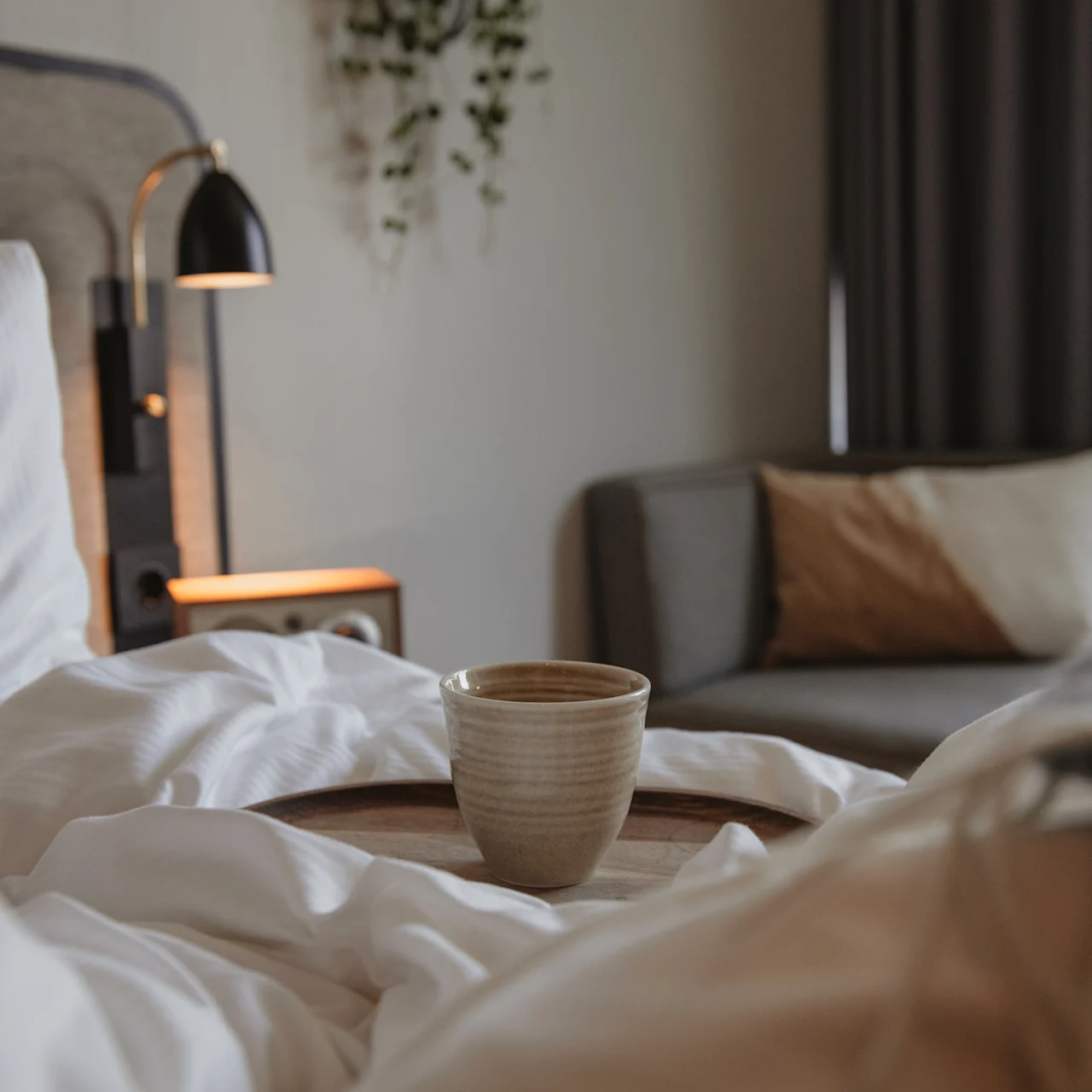A cup rests on a wooden tray atop a bed with white blankets. A modern lamp on a bedside table softly illuminates the room, which includes a couch with pillows.