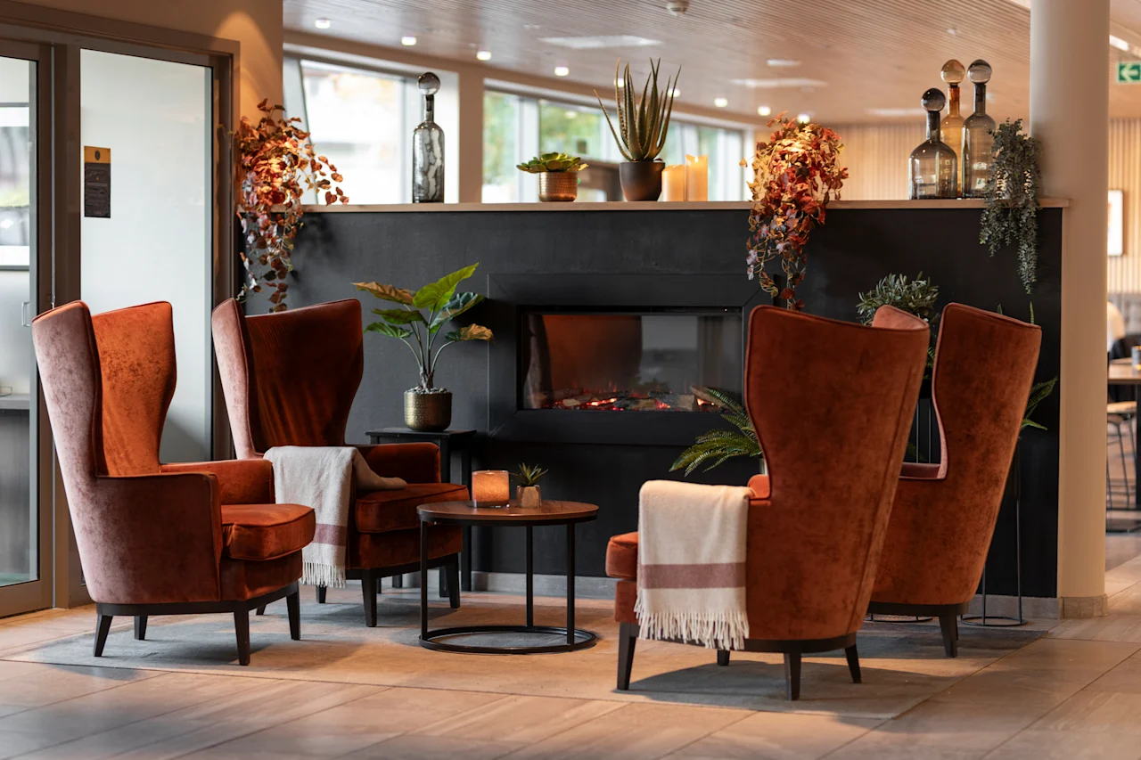 En hyggelig hotellounge med fire orange lænestole, en tændt pejs og dekorative planter, der indbyder til afslapning.