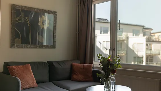En bekväm hotellvardagsrumsdel med en soffa, ett soffbord med blommor och en ljus fönsterutsikt över byggnader.