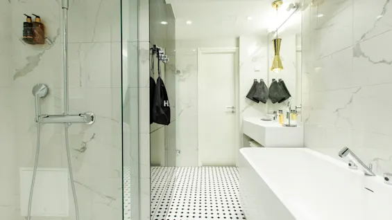 A modern bathroom features a glass shower, white bathtub, and marble walls; towels and toiletries adorn the vanity. The black and white tiled floor adds contrast.