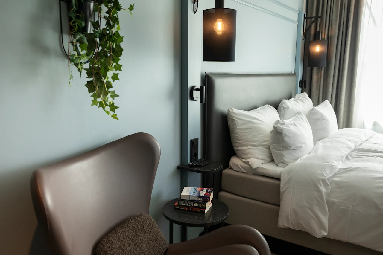 Et koselig hotellrom med seng, hvit sengetøy, komfortabel lenestol og hengende plante, opplyst av stilige vegglamper.