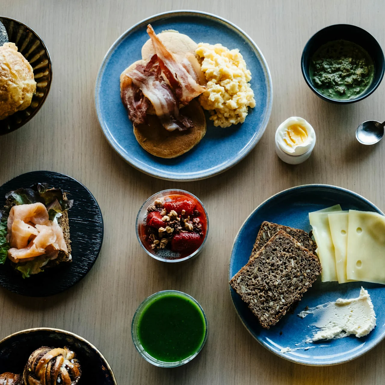 Pancakes with bacon and scrambled eggs on a blue plate, surrounded by various breakfast dishes like granola with strawberries, salad, bread with cheese, soup, and juice on a wooden table.