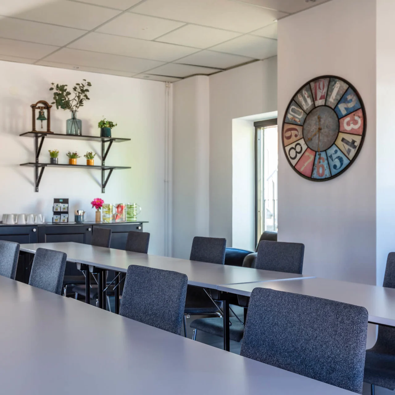 A modern conference room with a long table and chairs facing a kitchenette, decorations on the wall, and a large colorful clock.