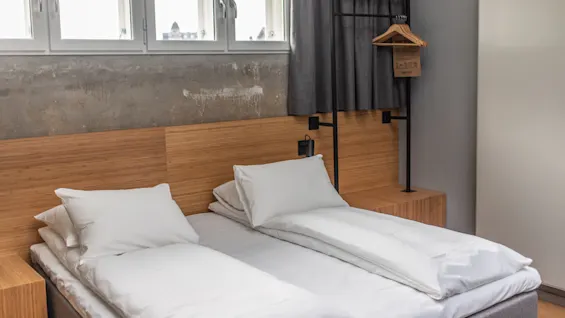 A double bed with white linens stands in a modern room with a wooden headboard. Above, small windows let in light. A clothes rack holds hangers beside the bed.