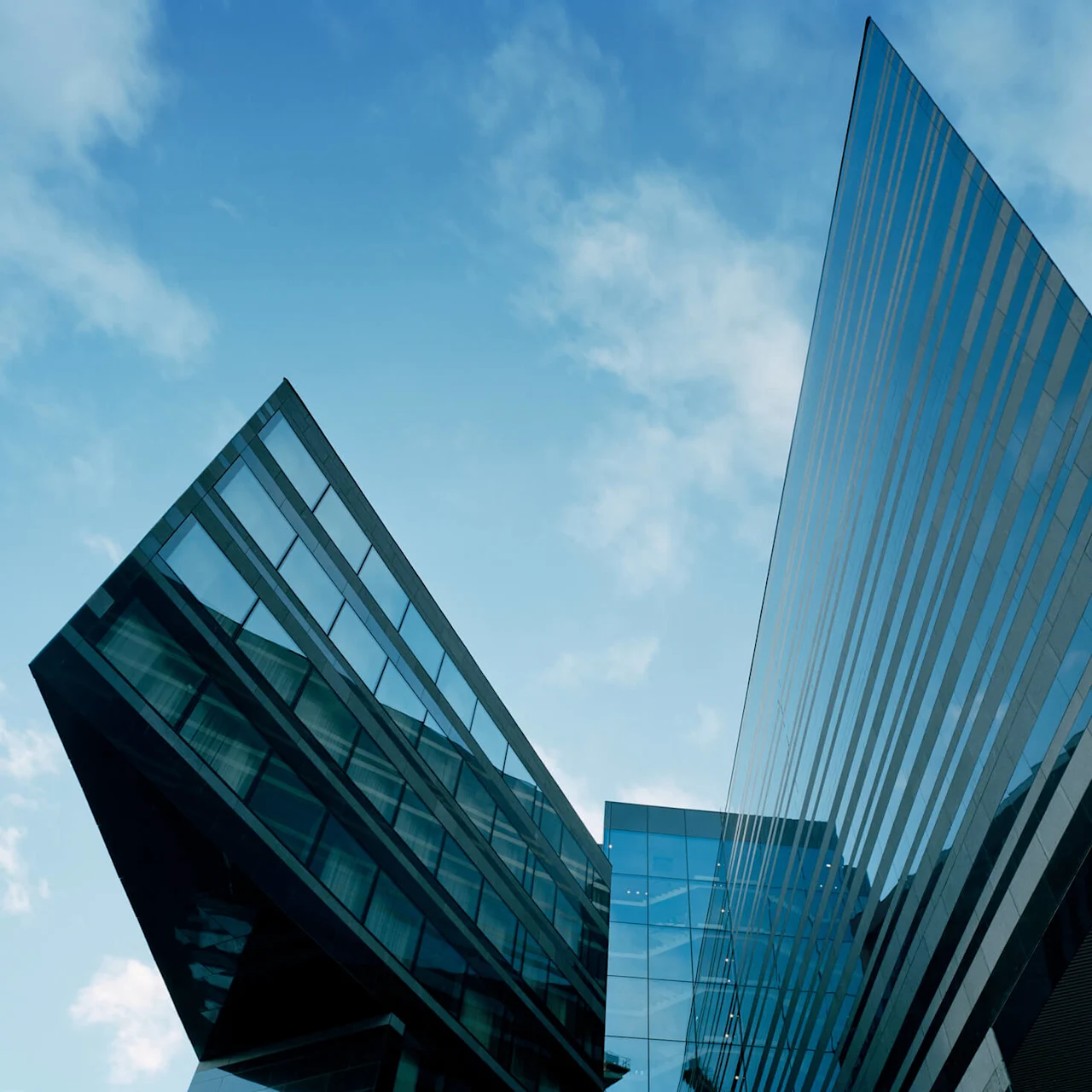 En modern, vinklad glasbyggnad sträcker sig uppåt och speglar den blå himlen med spridda moln. Den arkitektoniska designen har skarpa, korsande linjer och skapar en dynamisk urban estetik.