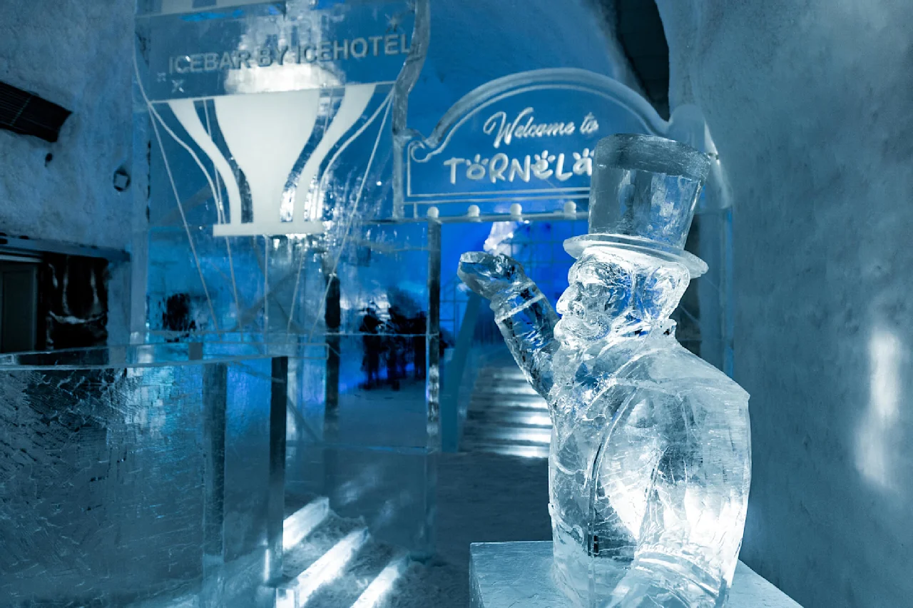 Isskulptur af en mand med høj hat, der løfter armen. Omgivelserne er et iset interiør med skilte med teksten "ICEBAR BY ICEHOTEL" og "Welcome to TORNEÄLV".