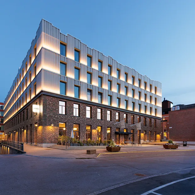A modern, multi-story building with illuminated upper sections and brick lower levels stands in an urban setting at dusk, surrounded by a paved area and neighboring structures.