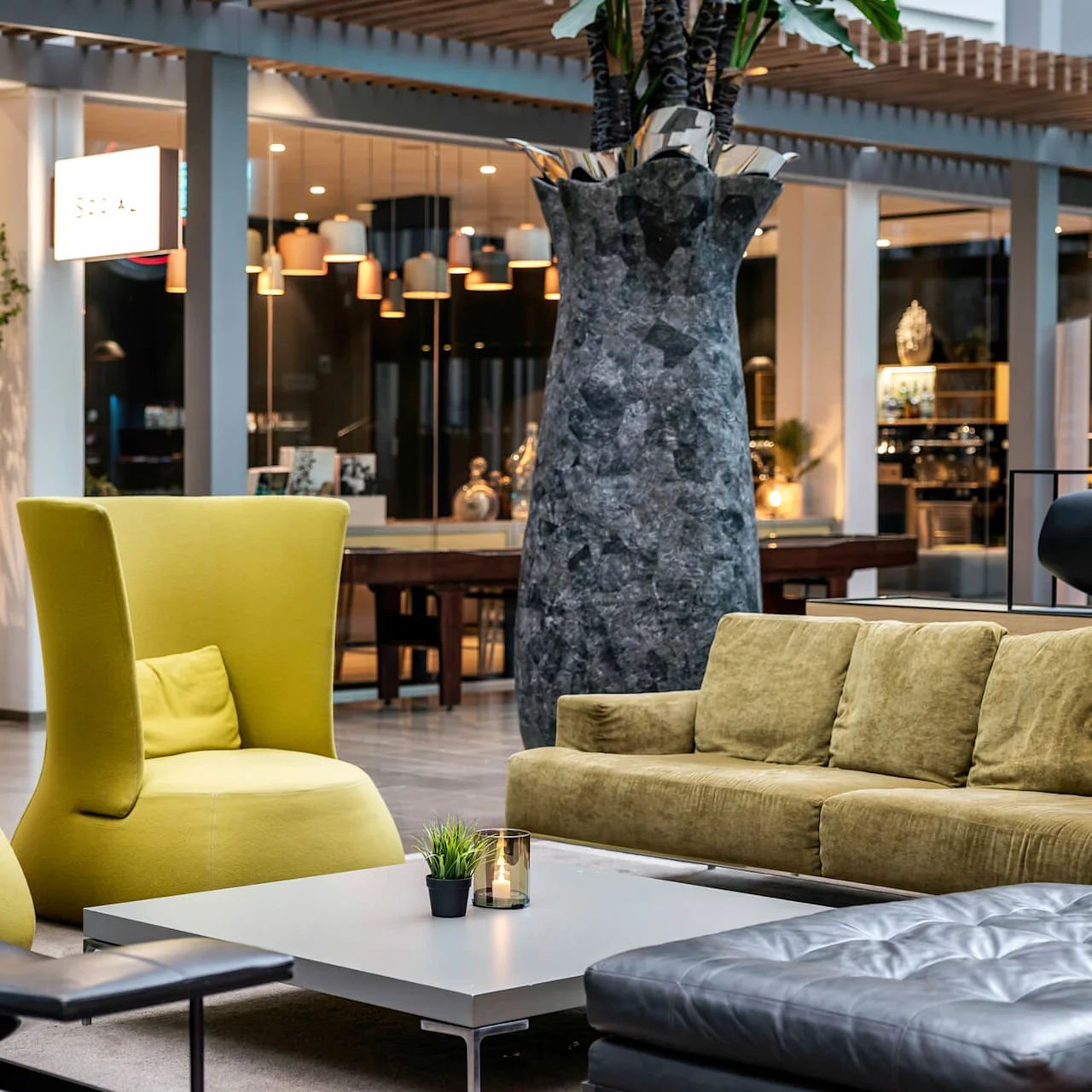 Gule lenestoler og en grønn sofa omgir et hvitt salongbord med et stearinlys og en plante. Under et stort tre tilfører lilla blomster farge til det moderne innemiljøet.