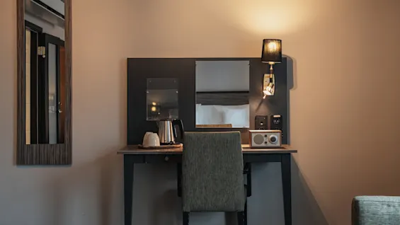 Hotellihuoneen työpöytä tuolilla, vedenkeittimellä, kupeilla ja radiolla, joka näyttää 0 0. Lämmin valaistus ja peilit täydentävät kutsuvan tilan.