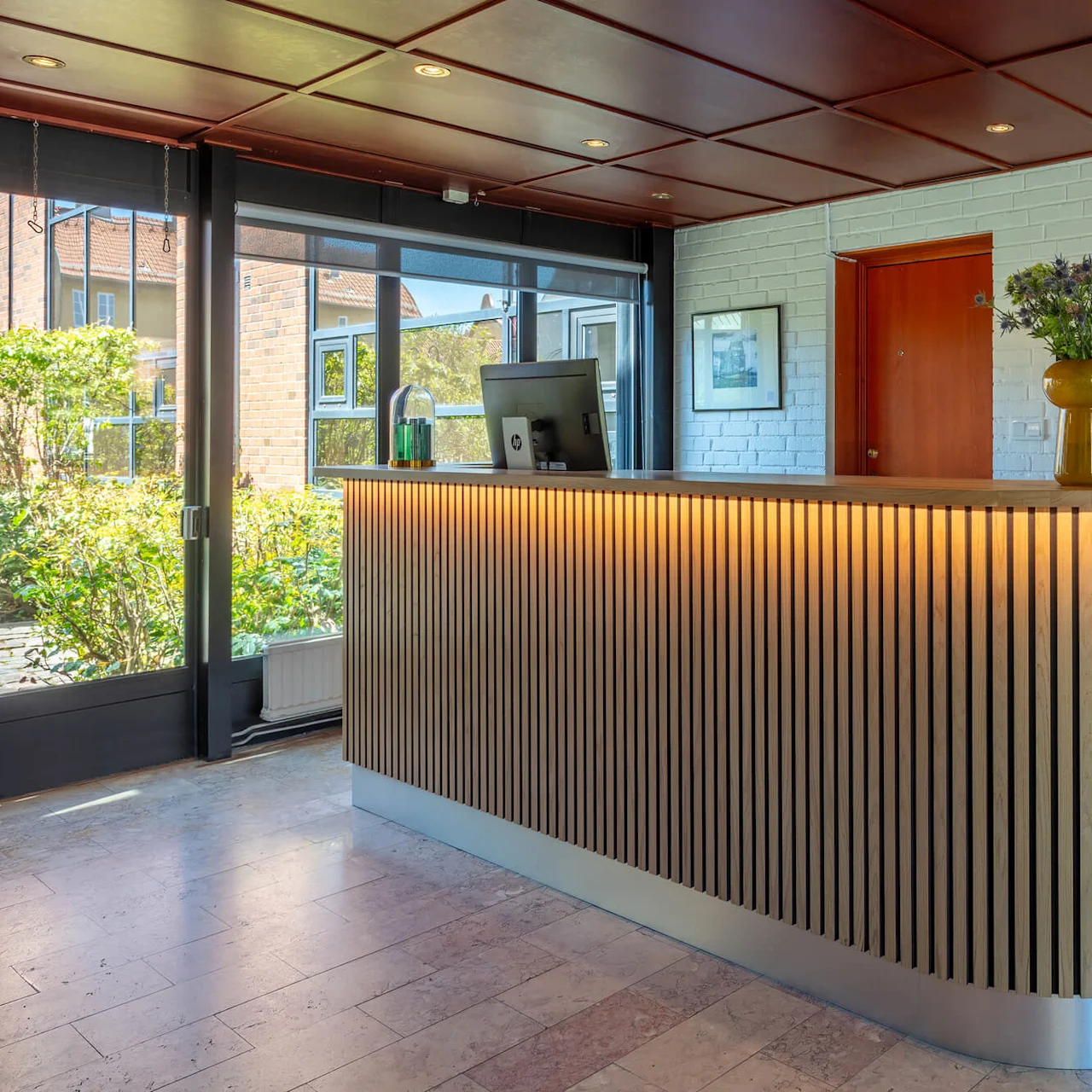 A curved reception desk with vertical slats, computers, and a flower vase, indoors with bright lighting and a view of greenery outside.