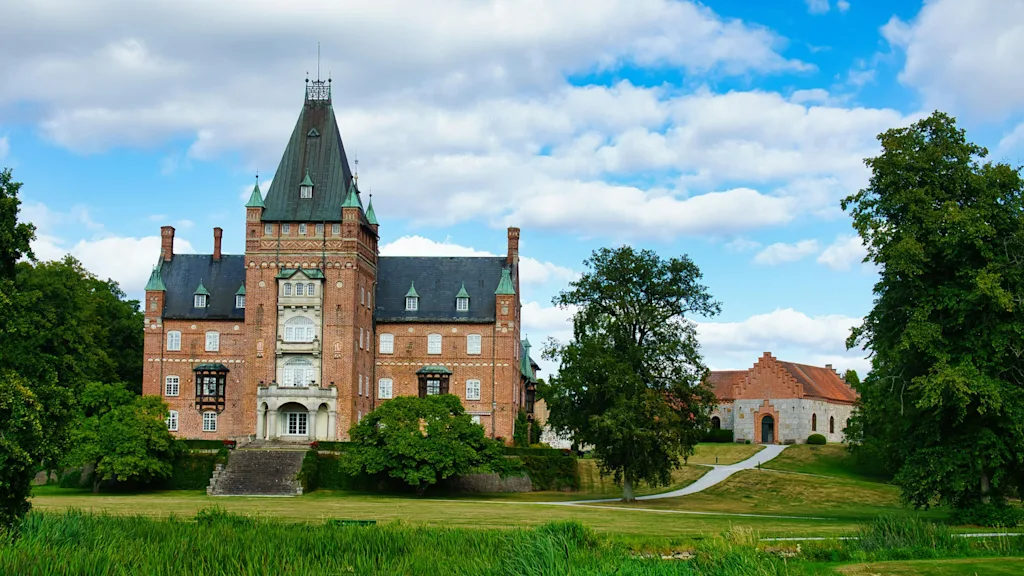 Et storslået murstensslot med et højt spir, omgivet af grønne træer og et frodigt græslandskab under en blå himmel.