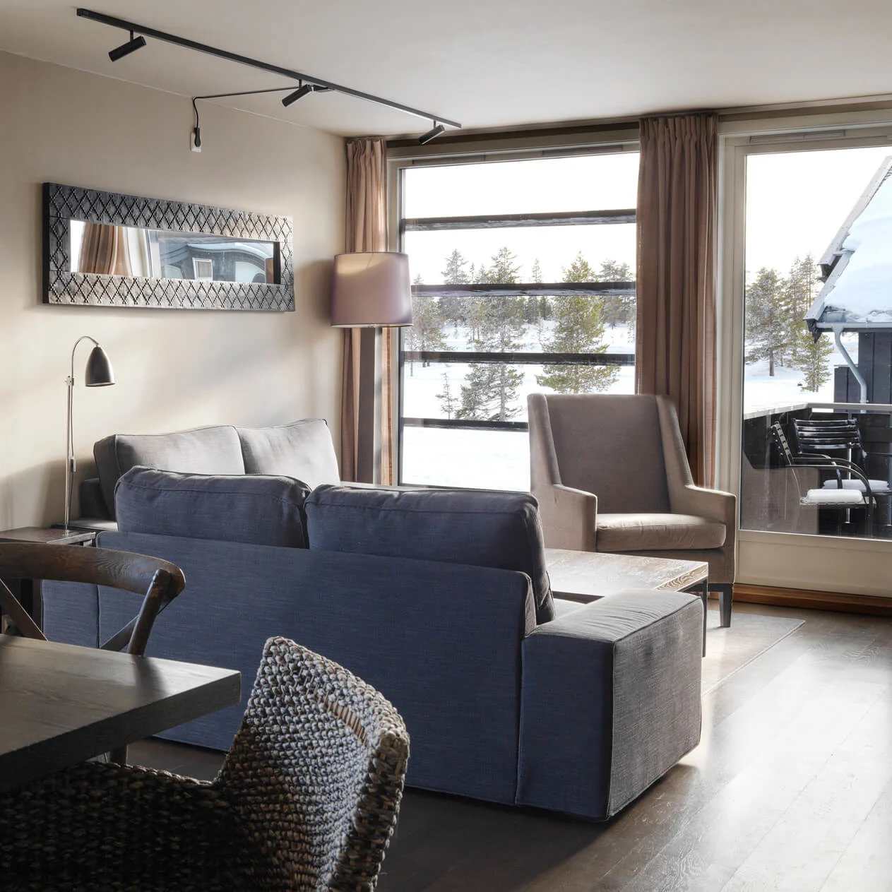 A modern living room features a gray sofa and armchair by large windows overlooking a snowy landscape, with a dark dining table and chairs nearby.