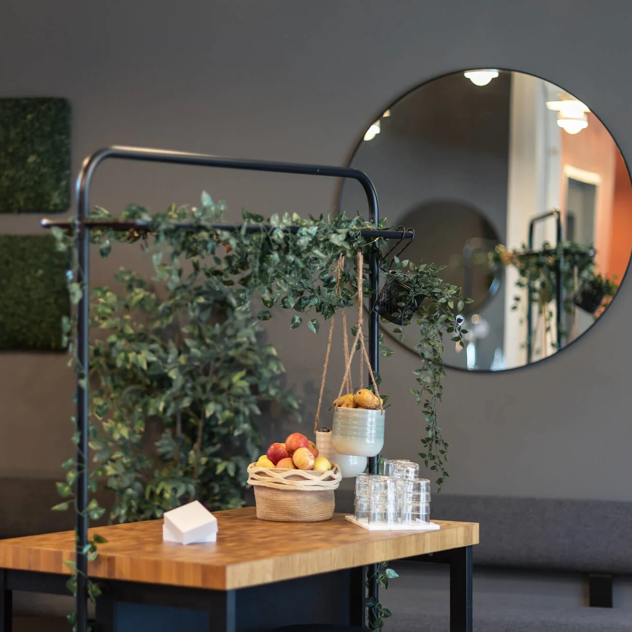 Wooden tables display baskets of fruit and hanging plant pots in a modern, greenery-decorated room with a circular mirror and rectangular moss art on the walls.