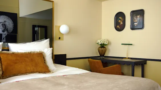 A neatly made bed with brown and white pillows stands beside a desk adorned with a vase of flowers, set against a wall with framed artwork and a decorative lamp.