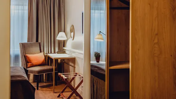 A cozy room with a chair, table, lamp, and a folding luggage rack, warm lighting, and a window with sheer curtains.