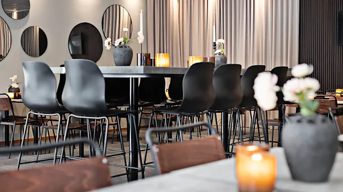 Svarte stoler omgir et høyt bord med vaser og lys. Sirkulære speil henger på en vegg ved siden av gulvlange gardiner, noe som skaper en moderne spiseatmosfære.