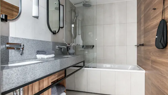 Et moderne badeværelse har en elegant dobbeltvask i granit med håndklæder og blomster. I nærheden er et brusehoved over et badekar omgivet af beige fliser og en træaccentvæg.