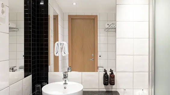 Ett modernt badrum med ett vitt handfat, en stor spegel och vita och svarta kaklade väggar, som reflekterar en trädörr.