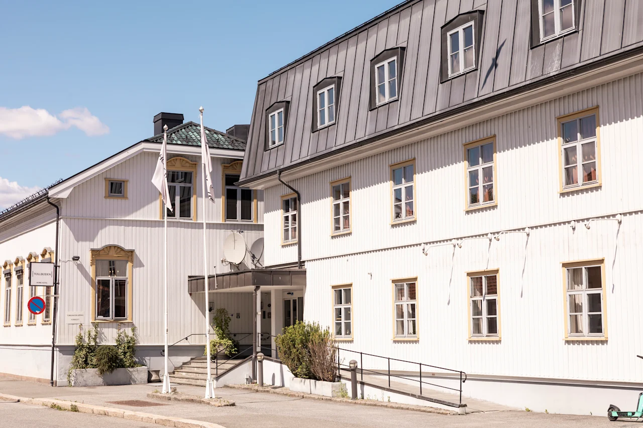 En vit, tvåvånings träbyggnad med många fönster och metalltak står på en solig gata under en klarblå himmel. Ingen text är synlig.