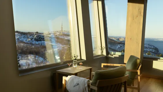 Et hyggeligt værelse med store vinduer og udsigt over sneklædte bjerge og hav. To stole, et bord med drinks og et tæppe med teksten WOOD.