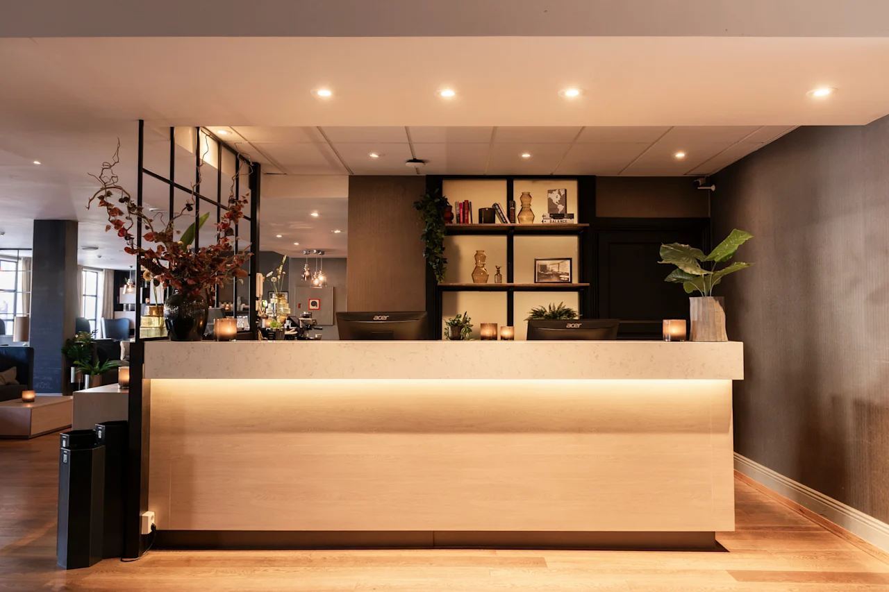 En varmt oplyst hotelreception med computerskærme, dekorative planter og hylder, der skaber en indbydende atmosfære.
