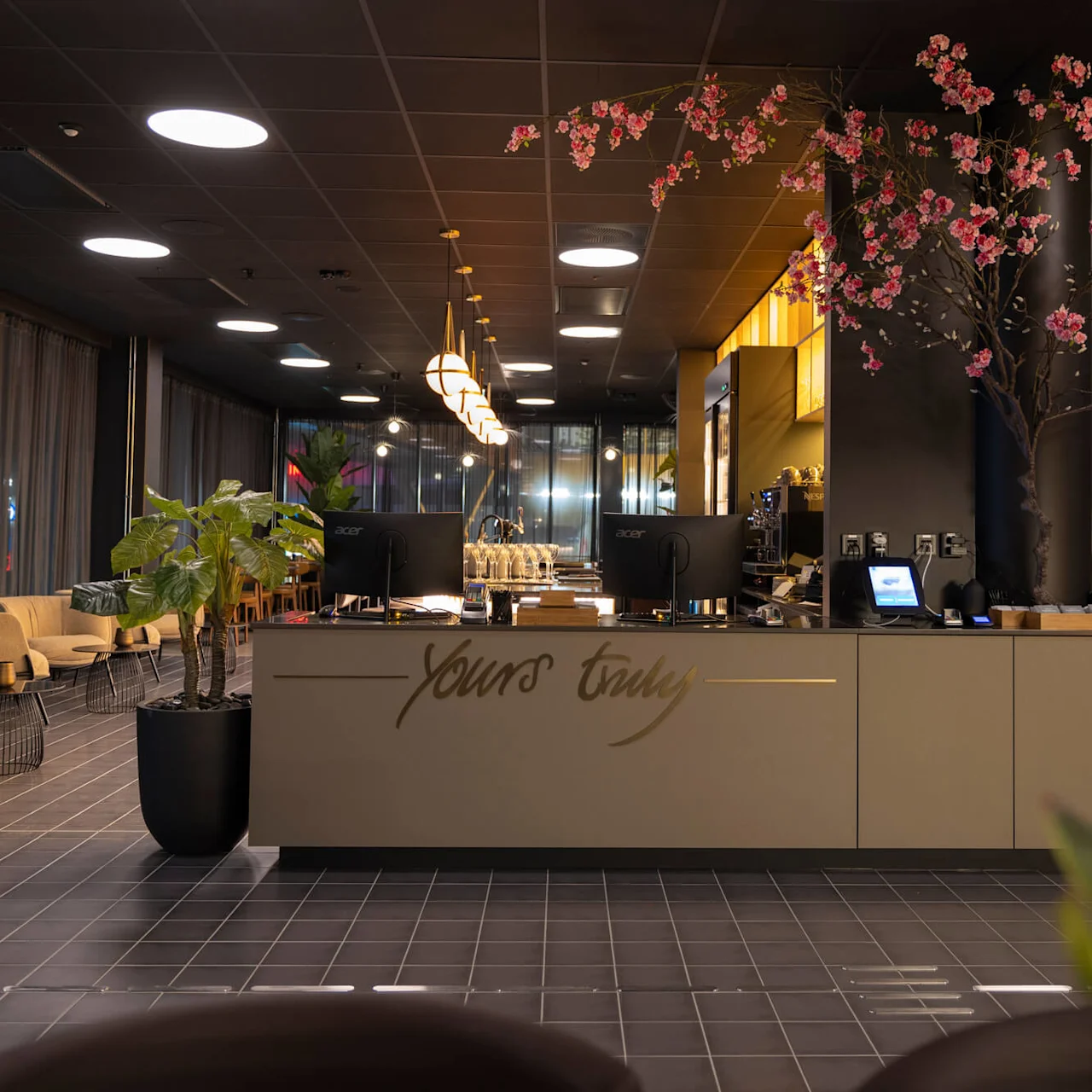 A modern cafe interior with a counter displaying "Yours truly" and blossoming tree, cozy seating in the background, and elegant lighting.