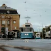 En blå sporvogn nærmer sig en station, glider langs skinnerne. Passagerer venter i nærheden, med en gammel murstensbygning og overskyet himmel i baggrunden.