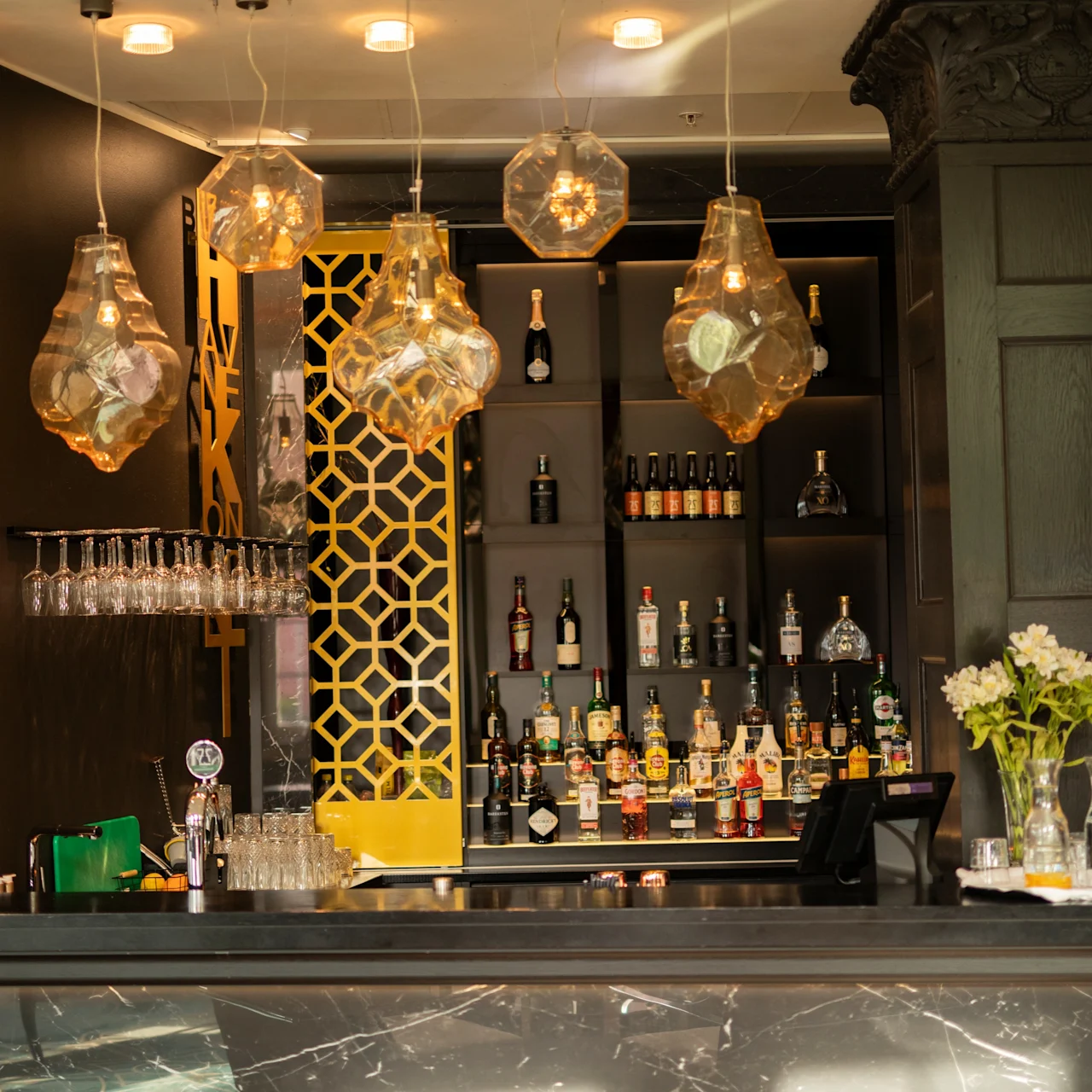 A stylish bar showcases hanging geometric lights, glassware, and diverse liquor bottles on shelves. Yellow geometric patterns and floral arrangements enhance the elegant hotel setting.