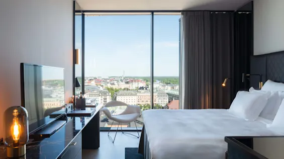 En pent oppredd seng vender mot et stort vindu med utsikt over bylandskap med bygninger og grøntområder under en klar himmel. Et skrivebord med lampe og stol står i nærheten.
