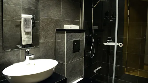 A modern bathroom with a white sink, towel rack, and toilet against black and gray tiles. A glass shower enclosure reflects the room.