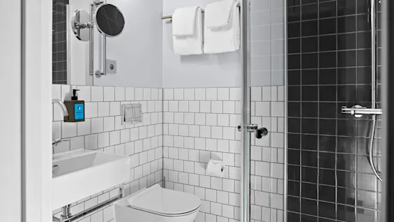 Et moderne bad med toalett, vask og dusj med glassvegger; veggene er kledd med hvite fliser. Håndklær henger på en stang. En blå såpepumpe er merket "SUE MUSE".