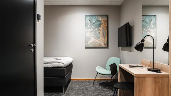 Ett litet sovrum innehåller en enkelsäng, ett skrivbord i trä med en svart lampa, mintgrön stol, väggmonterad TV, abstrakt konst och neutrala väggar.