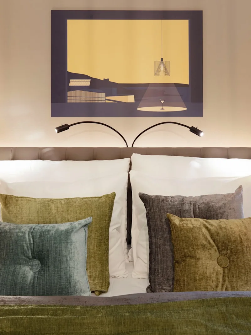 En prydligt bäddad säng har flera texturerade kuddar och en grön filt, flankeras av moderna lampor. Ovanför hänger ett minimalistiskt konstverk på en beige vägg i ett mjukt upplyst sovrum.