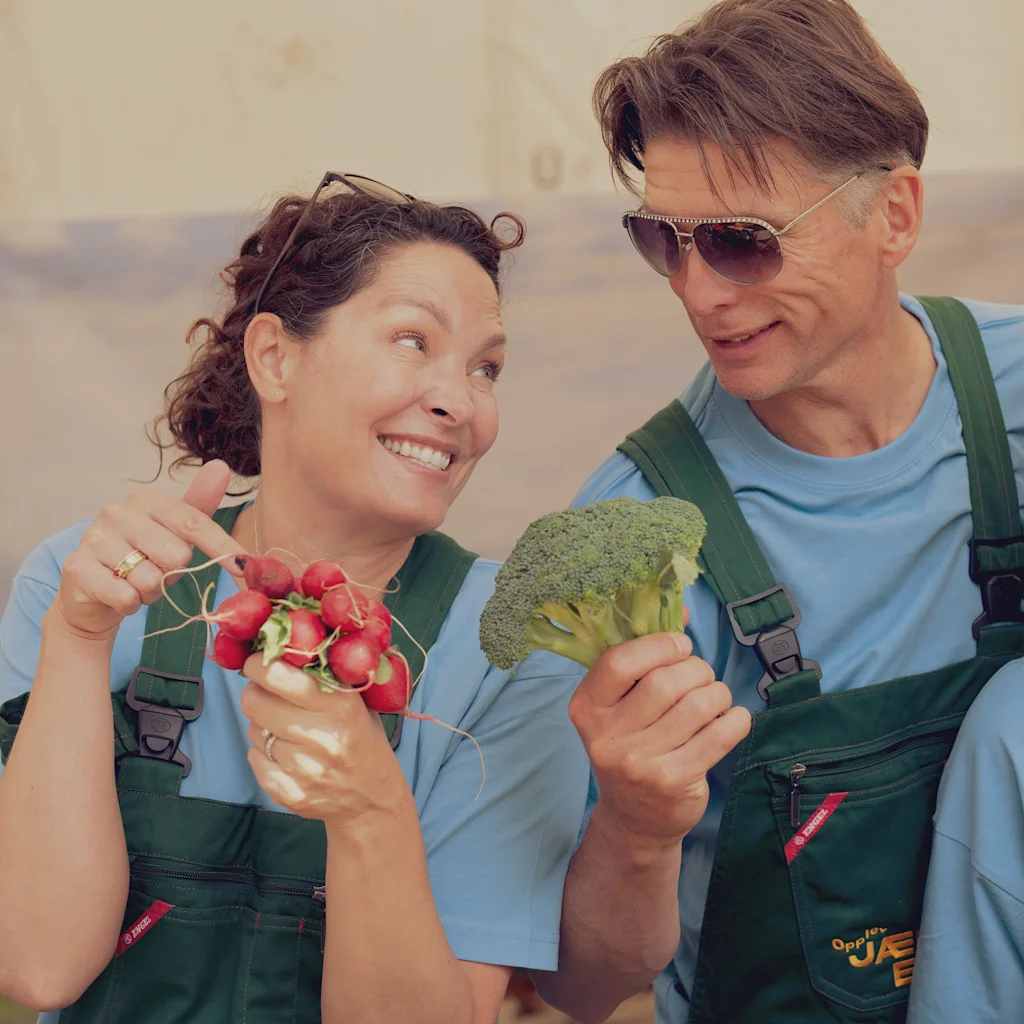 Kaksi haalareihin pukeutunutta ihmistä esittelee iloisesti vastapoimittuja retiisejä ja parsakaalia. Opplev JA