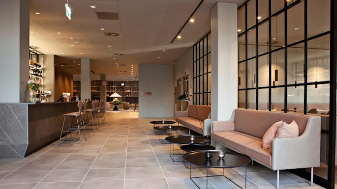 En moderne lounge har sofaer med puder ved siden af sorte sofaborde; en bar med høje stole ligger på den anden side. Blød belysning fylder det elegante, flisebelagte rum.