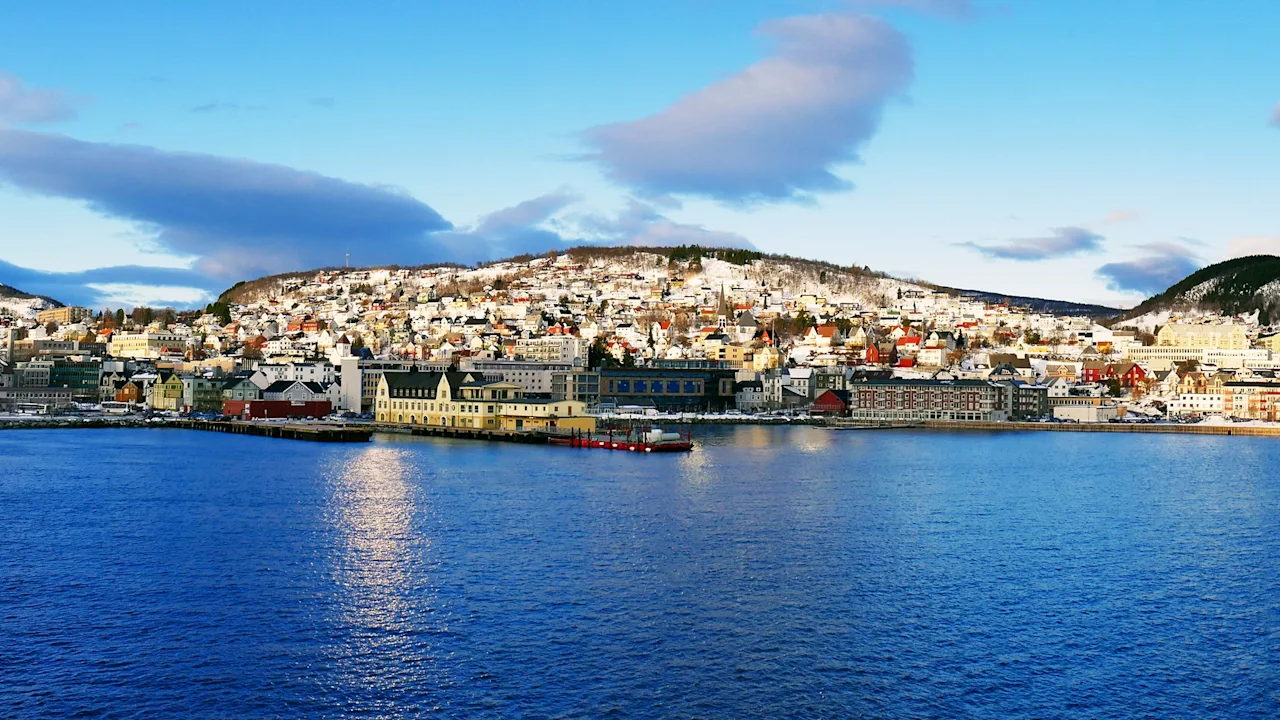 Buildings line a waterfront, resting below snow-dusted hills under a bright blue sky with scattered clouds. Calm water reflects the colorful structures.