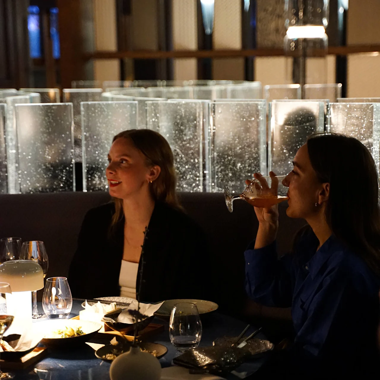 Kaksi naista istuu hämärästi valaistun pöydän ääressä, toinen juo lasista, toinen katsoo eteenpäin. Koristeelliset lasipaneelit ja himmeästi loistava lamppu luovat viihtyisän ravintolatunnelman.