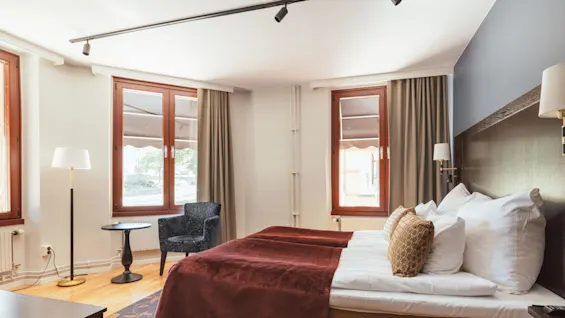 Et koselig soverom med en oppredd seng, nattbord, lampe, stol og åpne vinduer, som skaper en varm og innbydende atmosfære.