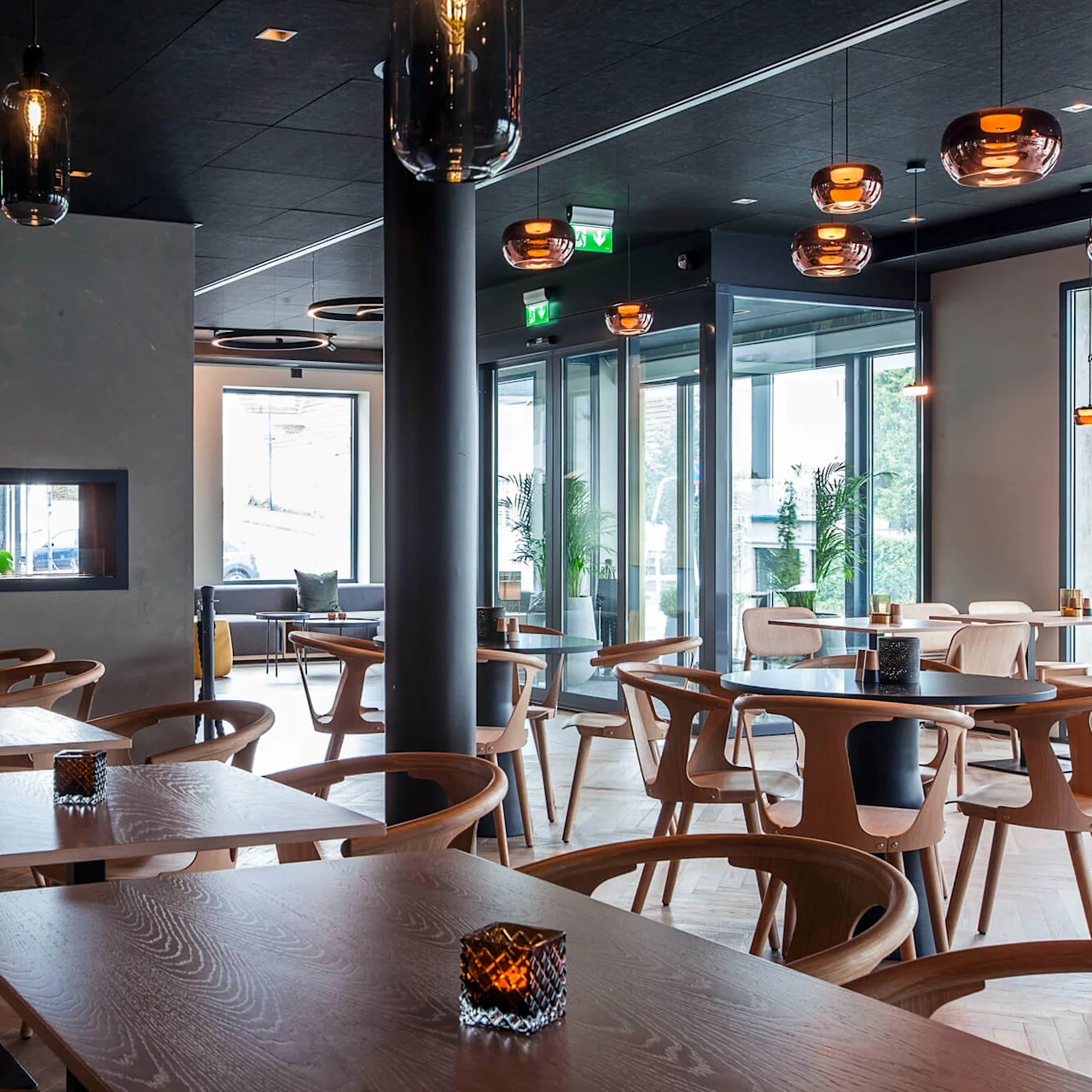 Bord og stoler er pent ordnet i en moderne restaurant med hengende lamper, en sentral peis og store vinduer som slipper inn naturlig lys.