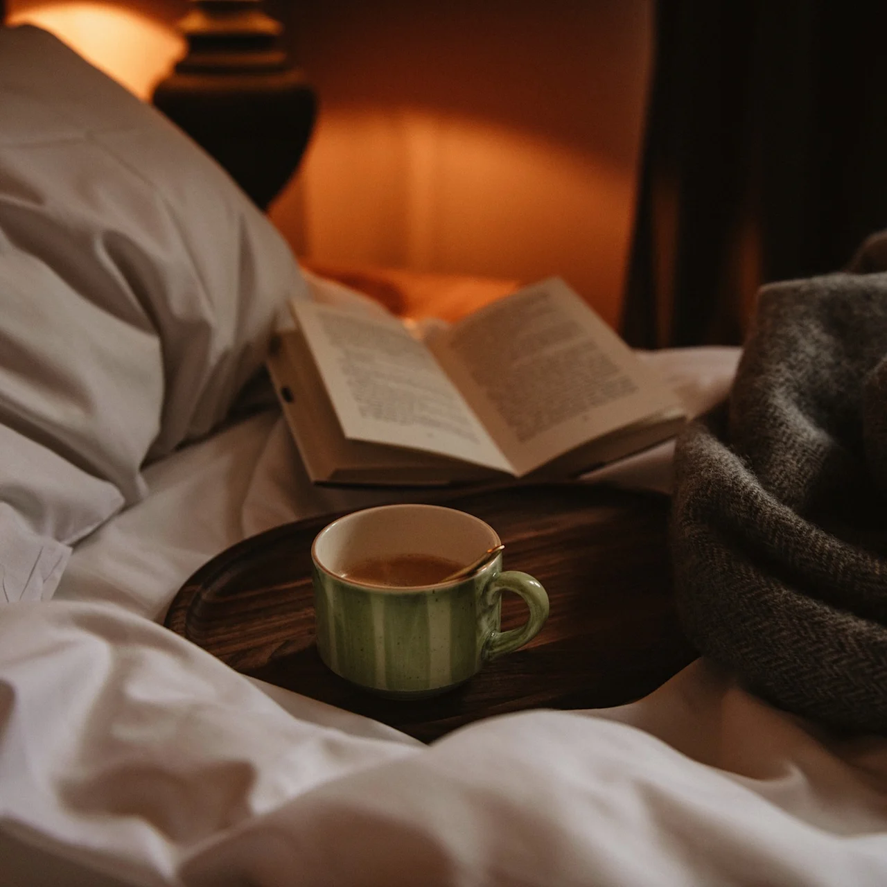 A green mug of coffee rests on a wooden tray atop a bed, accompanied by an open book and a cozy gray blanket nearby.