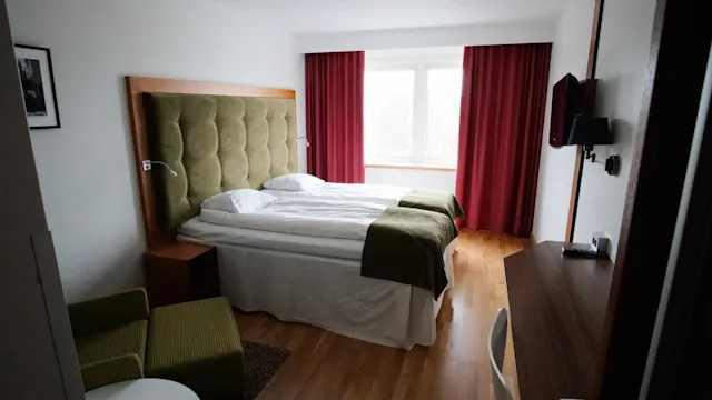 Siististi sijattu parisänky, jossa on vihreät tyynyt ja pehmustettu sängynpääty, seisoo hotellihuoneessa. Punaiset verhot kehystävät suurta ikkunaa, ja pieni pöytä on lähellä.