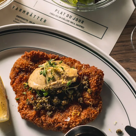 Breaded schnitzel topped with butter and capers, garnished with thyme, accompanied by a lemon wedge on a plate. On a wooden table with fries, red wine, and menu sheets.
