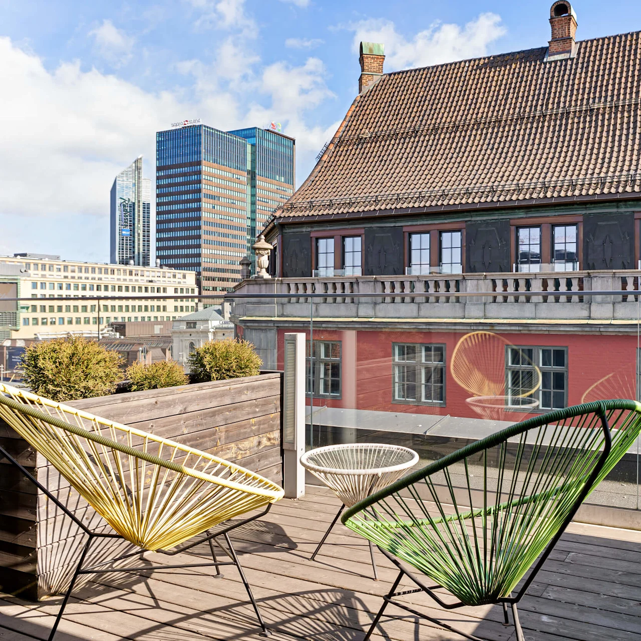 Two colorful chairs sit on a wooden deck, bordered by planters, overlooking a historic building with a contemporary cityscape in the background.