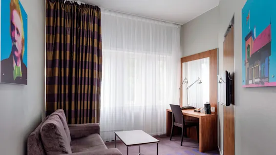 En lilla sofa vender mod et lille bord i et hotelværelse med stribede gardiner. Et skrivebord holder to lamper og en stol ved vinduet. Ingen synlig tekst.