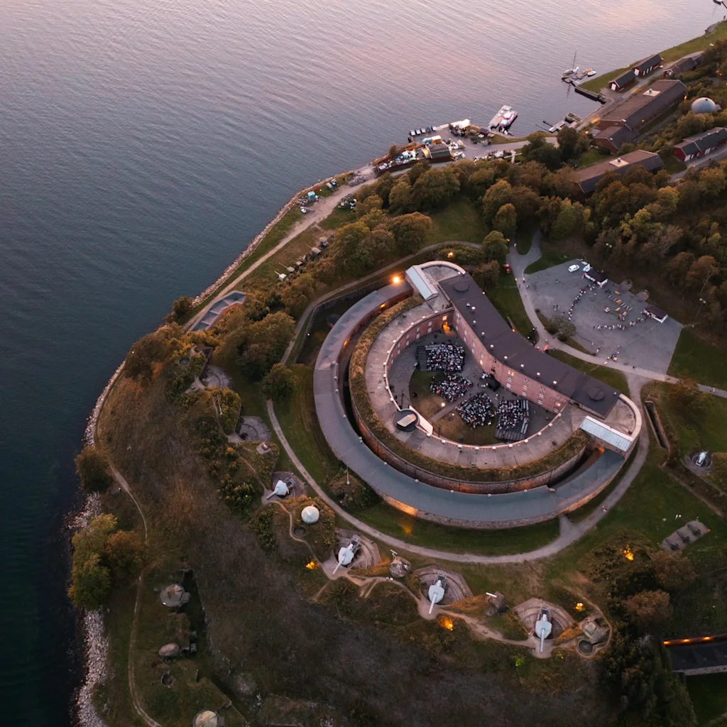 Luftfoto av et historisk kystfort med et stort utendørs arrangement i gårdsplassen, omgitt av trær og vann i skumringen.