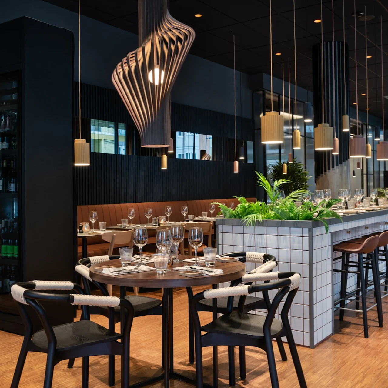 Elegante spisebord dekket med glass og bestikk, omgitt av moderne stoler, står i en elegant restaurant med hengende lys og frodig grønt, som skaper en sofistikert spiseatmosfære.