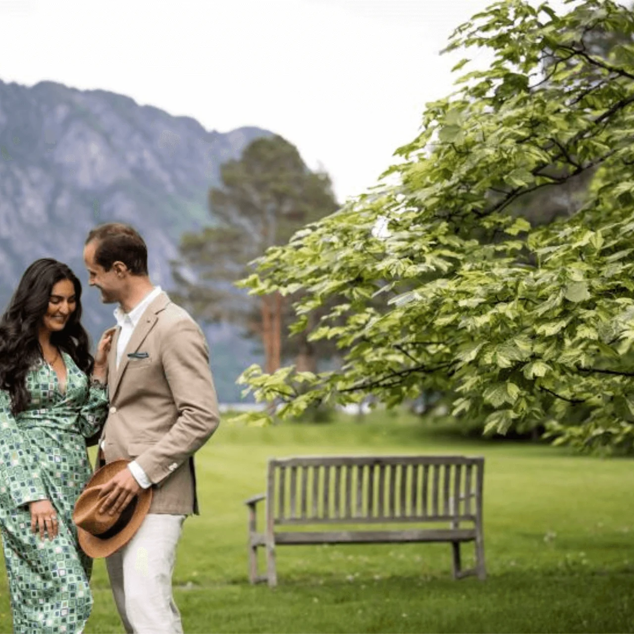 A couple stands closely on lush green grass, with the man holding a hat. They're surrounded by trees and mountains, with a wooden bench nearby.