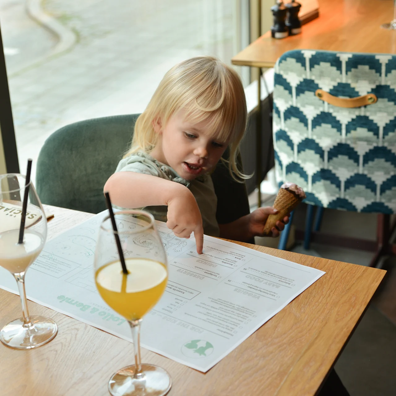 En barn peger på en menu, mens den holder en isvaffel i en restaurant. To drikkevarer med sugerør er på træbordet ved siden af en mønstret stol.