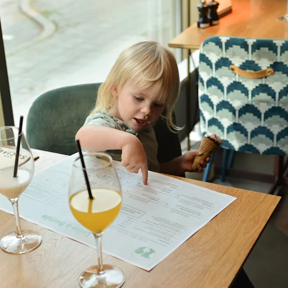 A child points at a menu while holding an ice cream cone in a restaurant. Two drinks with straws are on the wooden table, next to a patterned chair.