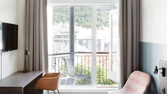 Et lyst hotelværelse med skrivebord, lampe, stole og et vægmonteret TV. En åben dør fører til en balkon med udsigt over hustage og grønne bakker.